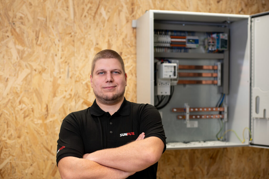 Mann im SunFuse-Poloshirt vor offenem PV-Sammelschrank, rechts Schrank mit Kupferschienen und Bauteilen