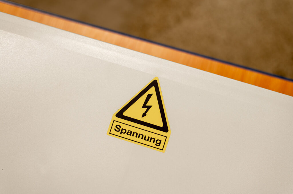 Gelber Warnaufkleber mit Blitzsymbol und Text „Spannung“ mittig auf weißem Gehäusedeckel, oben Holzrand