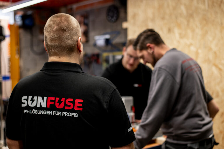 Mann mit Rücken zur Kamera, Shirttext „SUNFUSE PV-LÖSUNGEN FÜR PROFIS“, rechts zwei Personen am Arbeitstisch