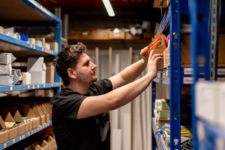 Mann greift im Lager zwischen blauen Regalen nach orangefarbenen Kabeln, links Kartons und Bauteile