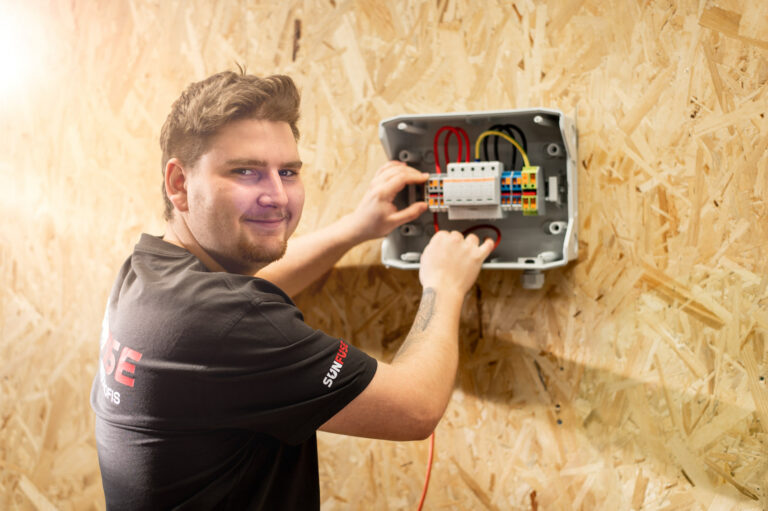 Monteur vor geöffneter PV-Box an OSB-Wand, links Person, rechts verdrahtete Klemmen und Modul, Text SUNFUSE