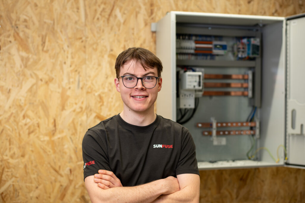 Mann mit SunFuse-Shirt vor Holzwand, rechts geöffneter PV-Sammelschrank mit elektrischen Komponenten und Kupferschienen