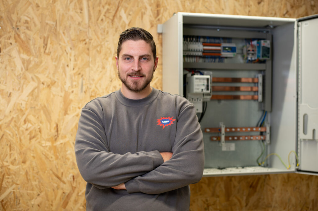 Mann mit verschränkten Armen vor offenem PV-Sammelschrank; Pullover mit Logo-Text CHIP, rechts Schrank mit Komponenten