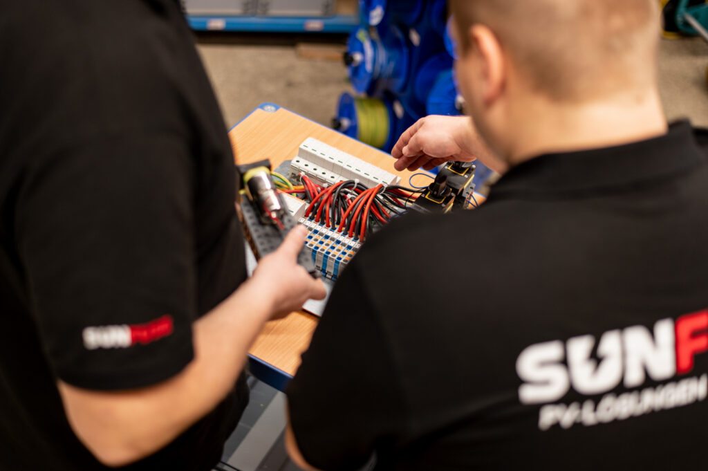 Zwei Personen in SunFuse-Shirts montieren am Tisch PV-Box mit PushIN-Klemmen, roten Kabeln und Schaltern.