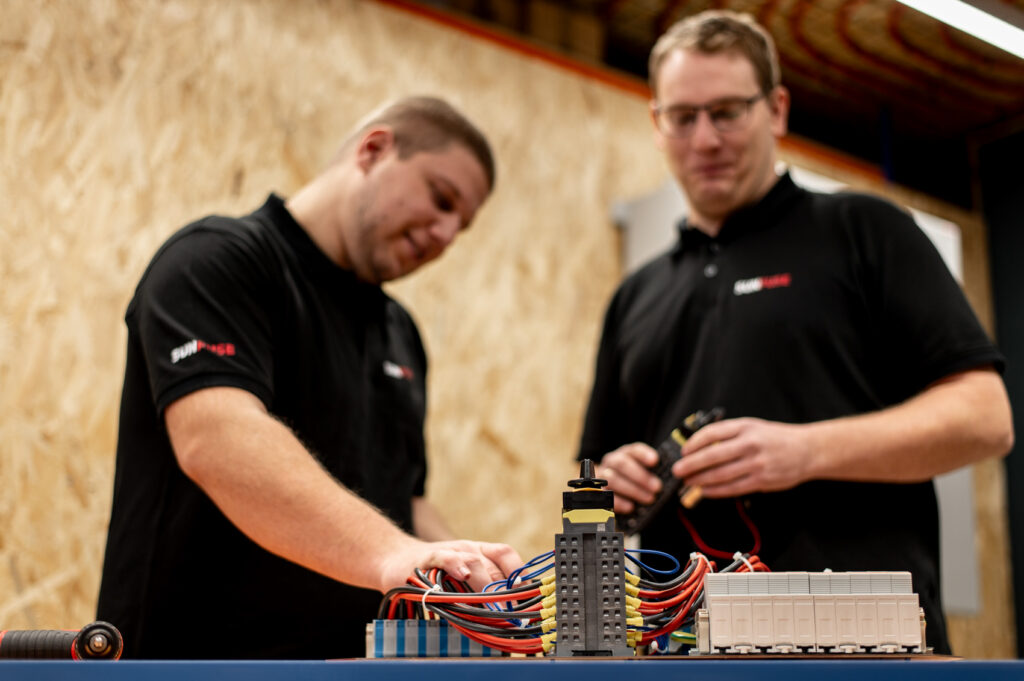 Zwei Mitarbeiter in SUNFUSE-Shirts montieren Werkbank, vorne Kabel und Klemmenblock, rechts weiße Anschlussmodule.