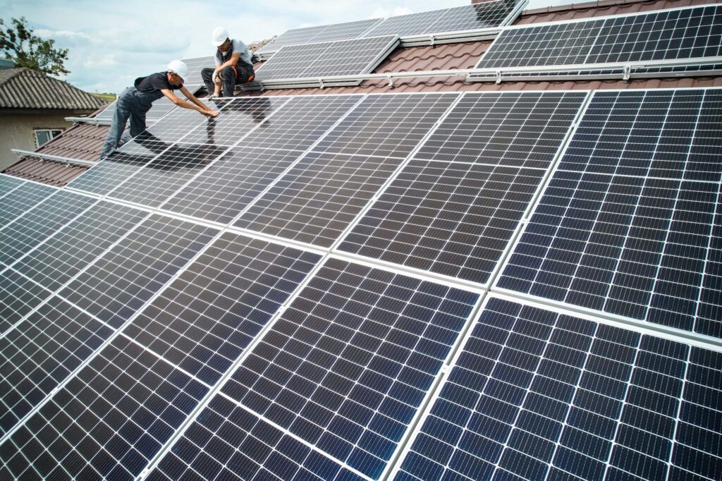Zwei Elektriker montieren Solarmodule auf Schrägdach; links stehend, oben kniend zwischen PV-Modulen.